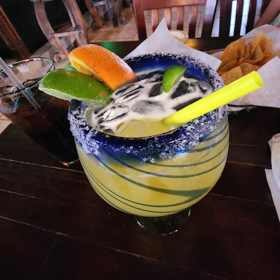 A large margarita in a blue-rimmed glass with lime and orange slices, next to a dark drink.