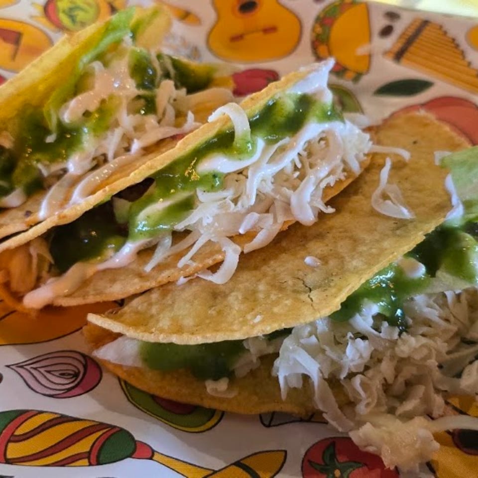 Two crispy tacos with green sauce, white sauce, and shredded cabbage on a colorful patterned paper.