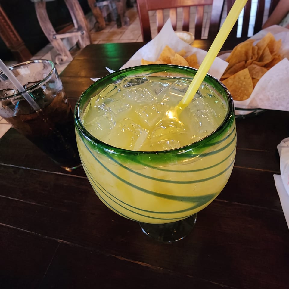 A large margarita with a yellow straw and a glass of soda on a dark wooden table.